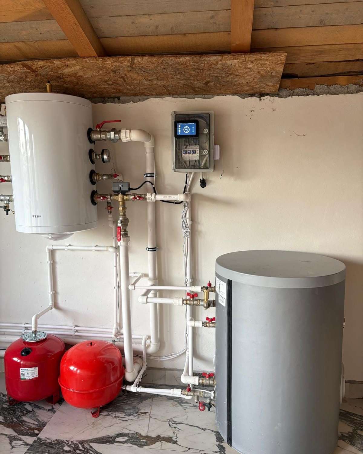 Cameră tehnică cu boiler alb, vas tampon gri, vase de expansiune roșii și trasee de țeavă albă
