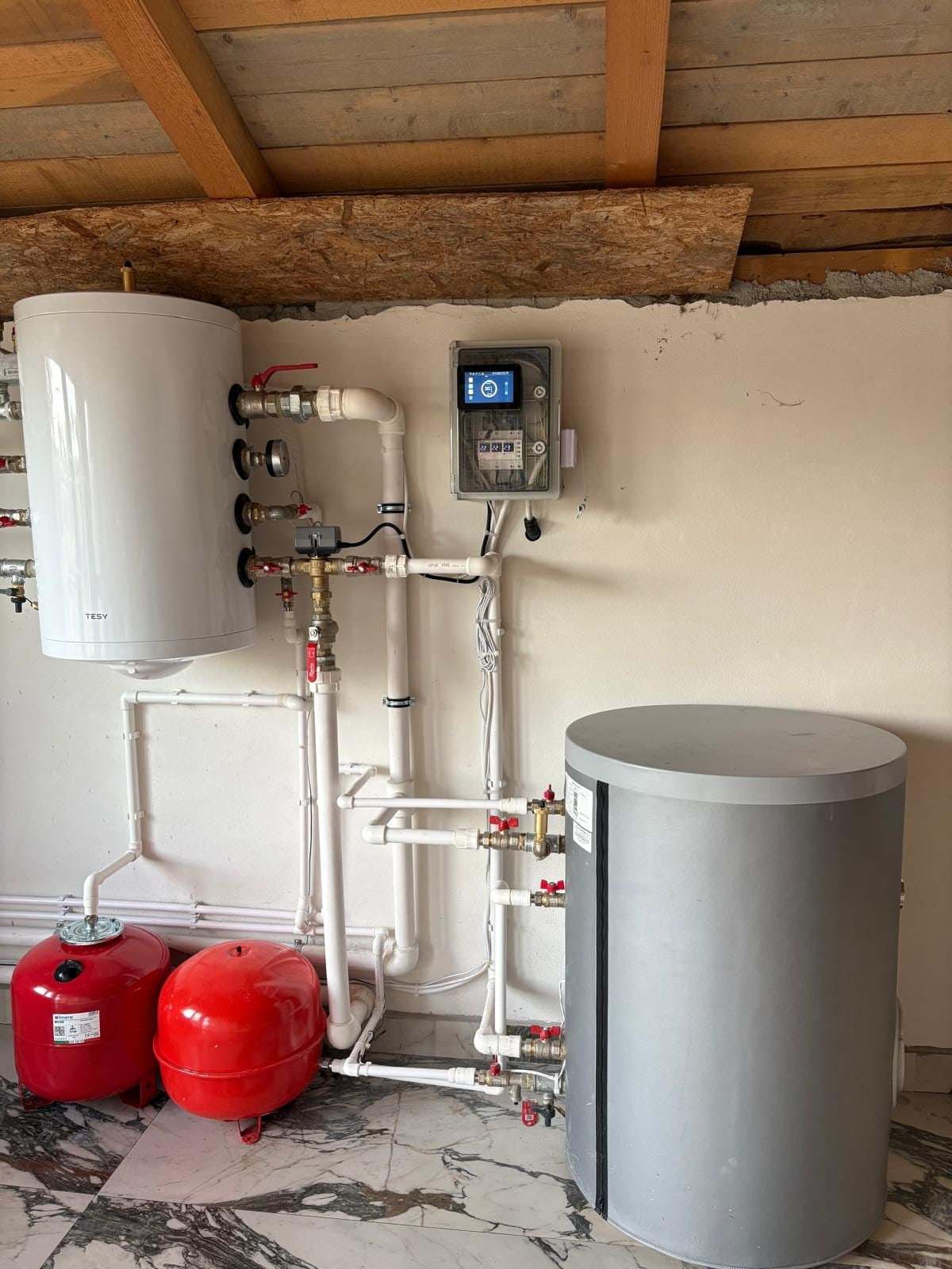 Cameră tehnică cu boiler alb, vas tampon gri, vase de expansiune roșii și trasee de țeavă albă