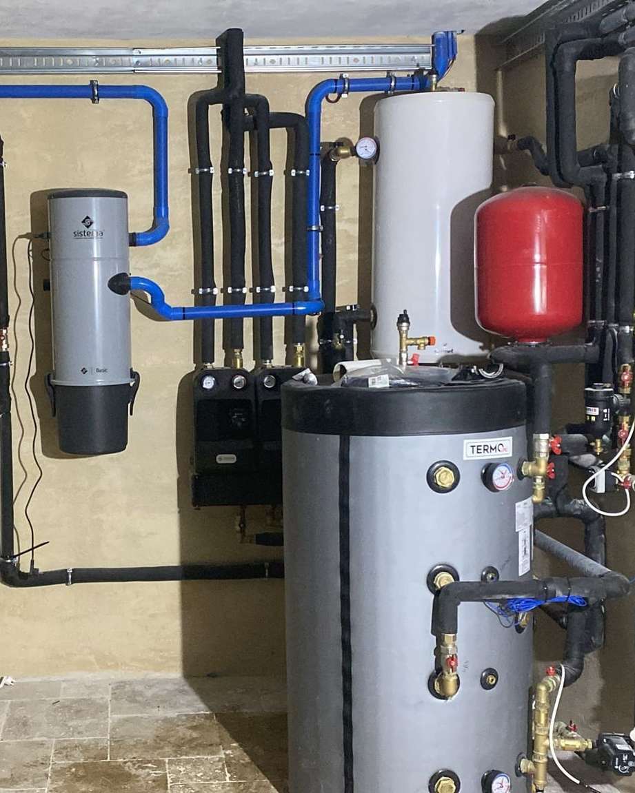 Cameră tehnică cu rezervor gri, boiler alb, vas de expansiune roșu și conducte izolate pe perete