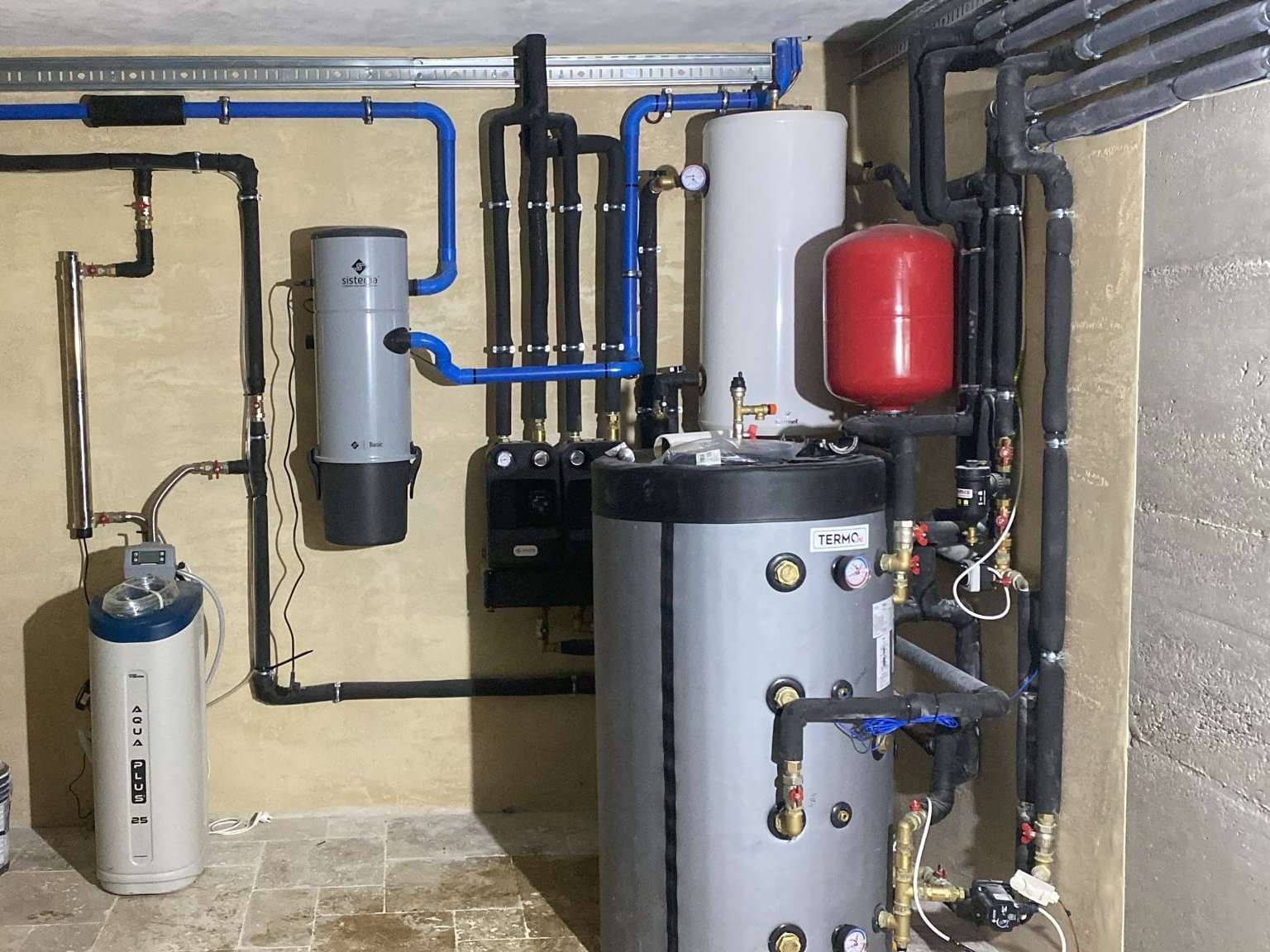 Cameră tehnică cu rezervor gri, boiler alb, vas de expansiune roșu și conducte izolate pe perete
