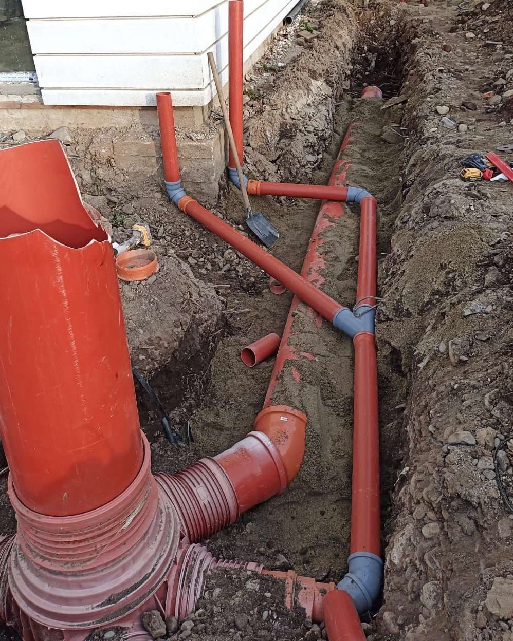 Tranșee lângă o casă, cu țevi portocalii de canalizare din PVC, ramificații și un cămin