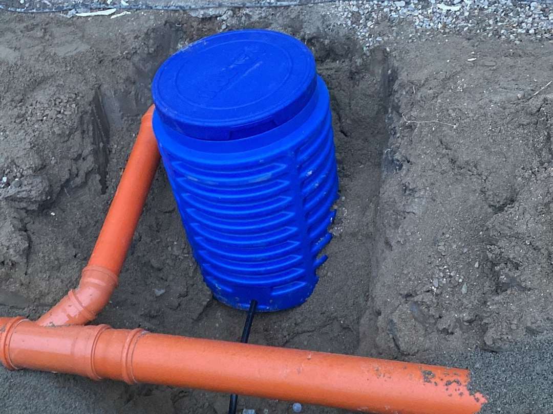 Cămin albastru de apometru într-o groapă, cu țeava portocalie de canalizare și conducta neagră de apă